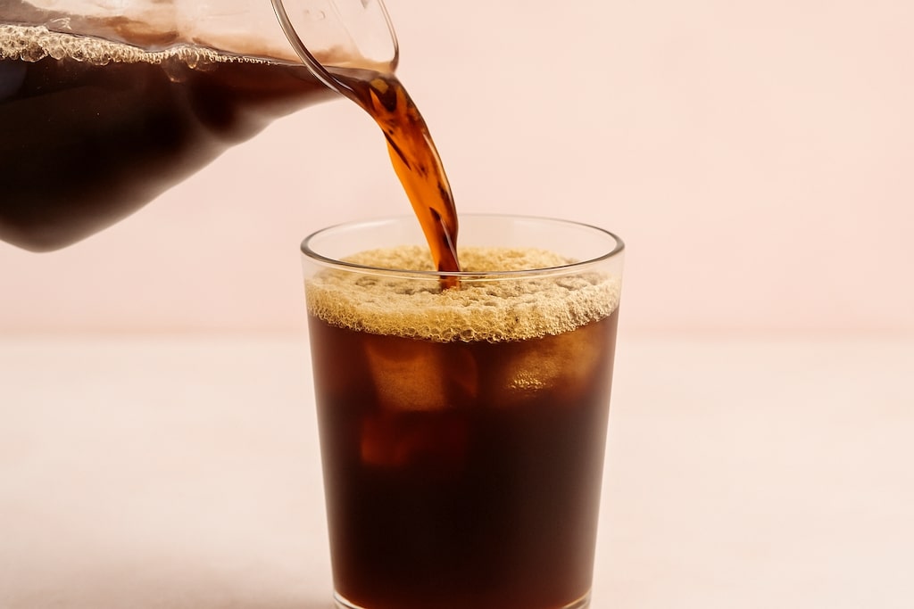 Cold Brew wird aus Kanne in Glas gegossen, auf rosa Hintergrund