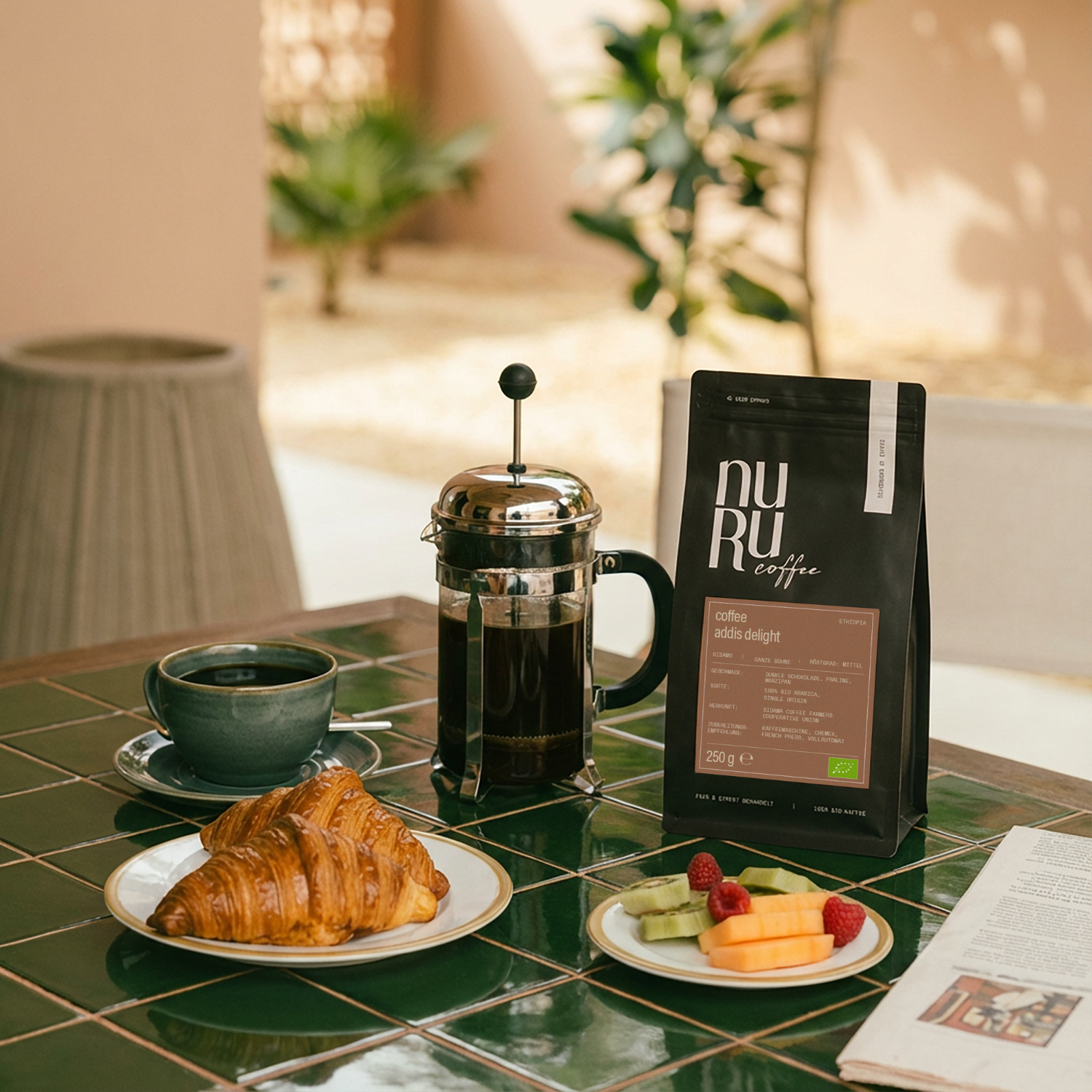 BIo Kaffee von nuruCoffee mit French Press, Teller mit zwei Croissants und Tasse mit schwarzem Kaffee auf Tisch mit grünen Fliesen