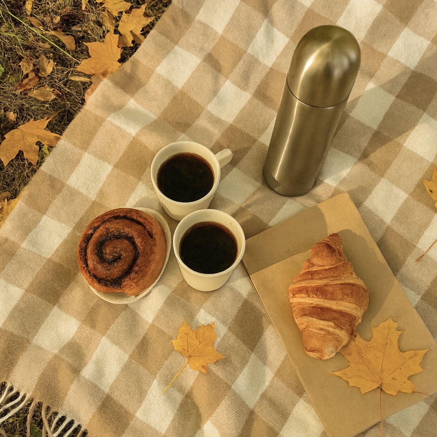 Croissant, Thermoskanne, zwei Tassen Kaffee und Zimtschnecke auf karierter Tischdecke