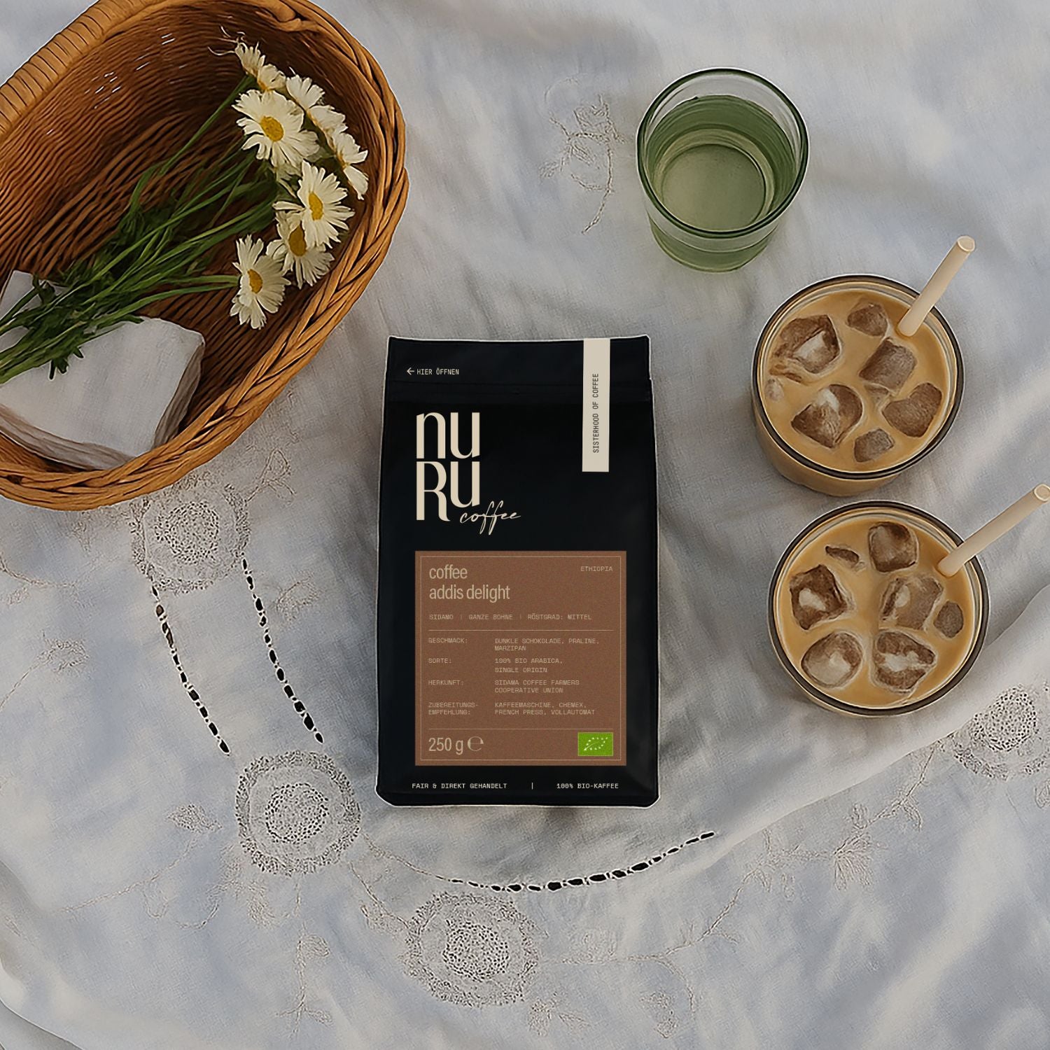 nuruCoffee Kaffeepackung auf weißer Picknickdecke mit Blumen und zwei Gläsern mit Eiswürfeln und Kaffee